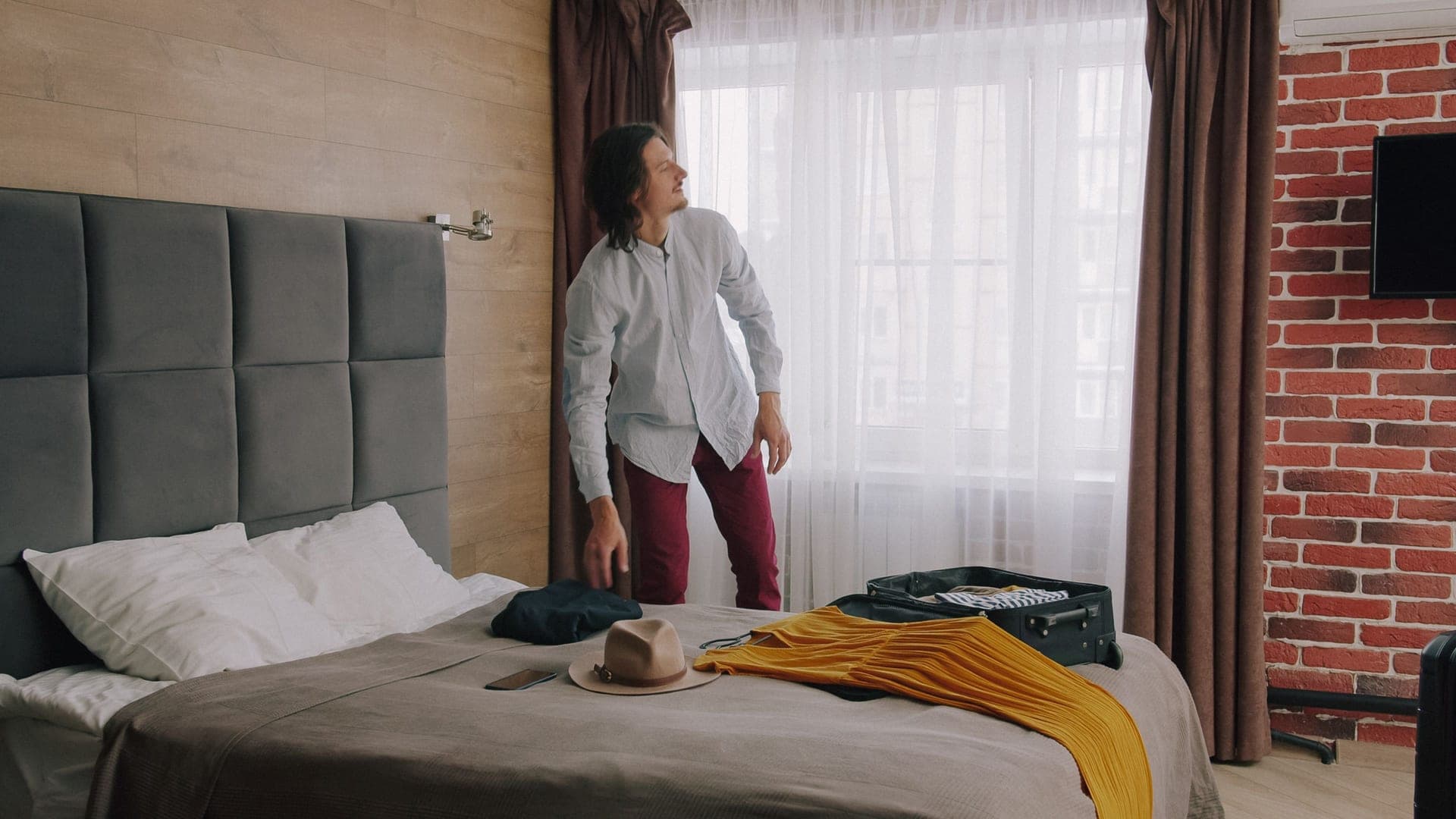 A hotel room with a made bed, a window and an open suitcase on the floor